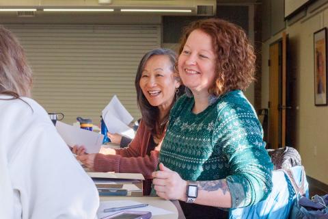 Two women sitting at a table smile and look to someone blurred in the lower left corner