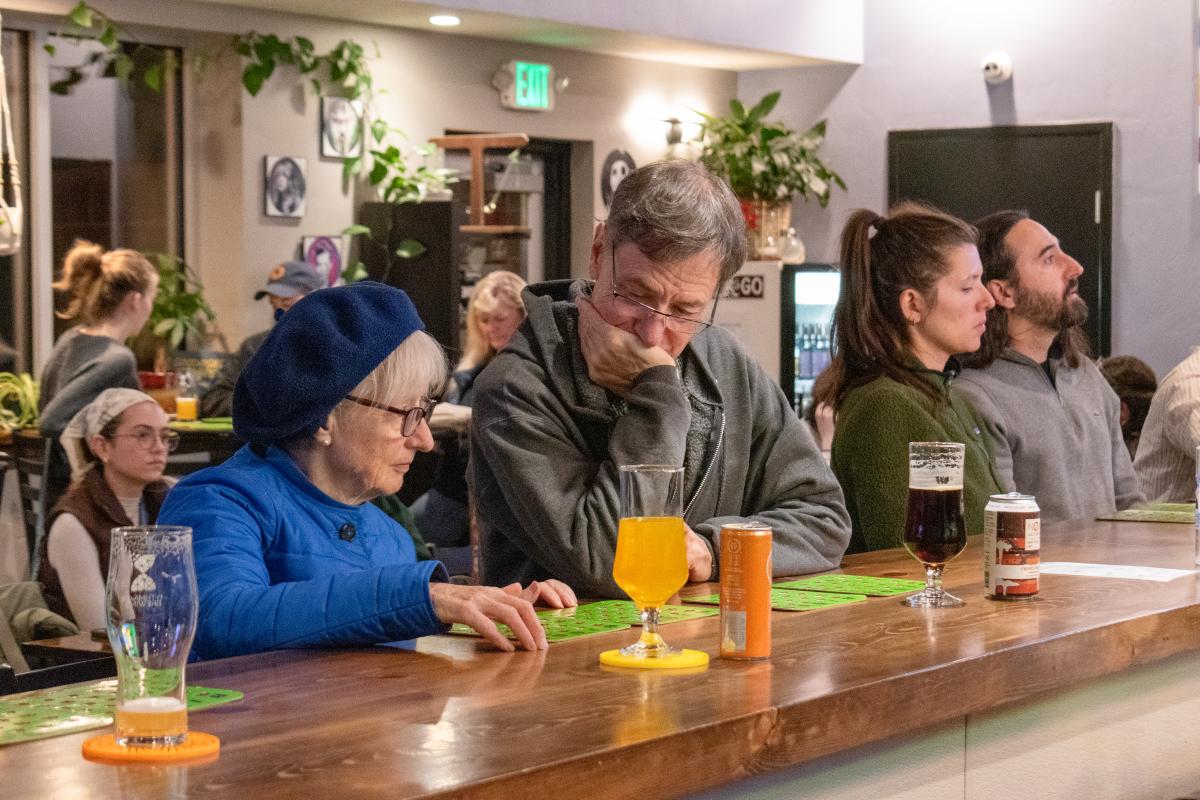 Older Couple Plays bingo at brewery bar. Beers sit in front of them and other patrons play in the background.