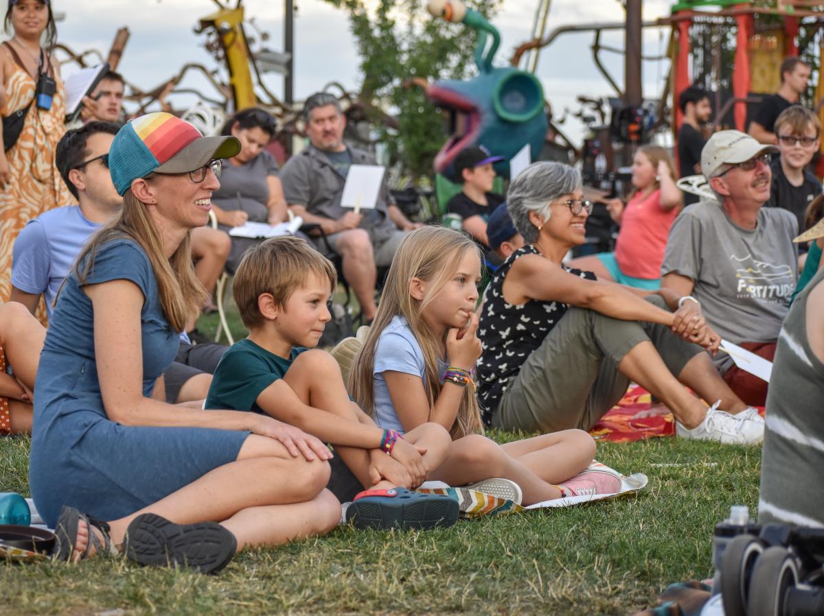 Family sits on grass for outdoor performance while others sit in the background.